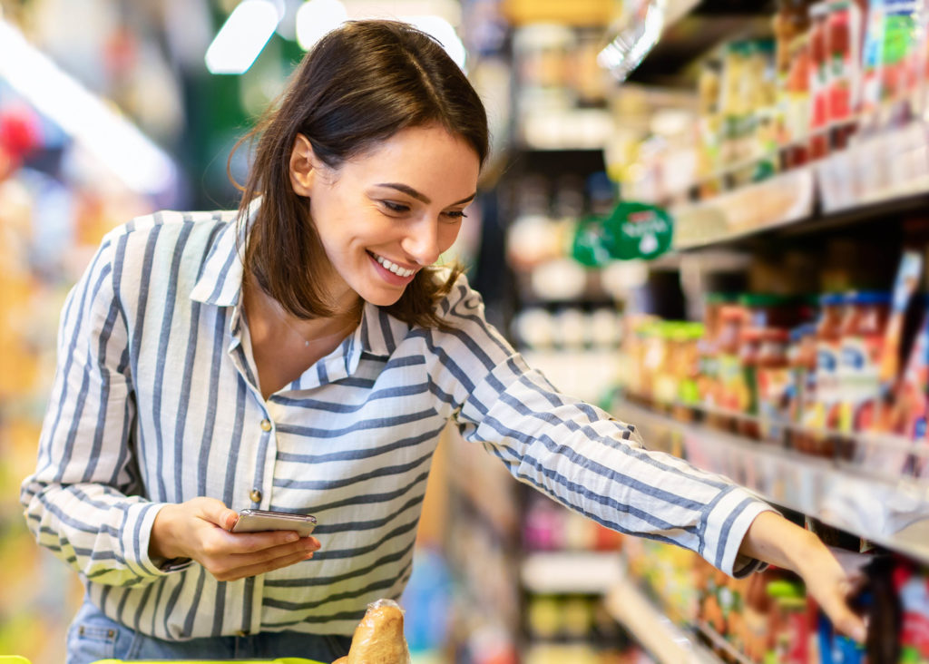 Shopping. Young Smiling Woman Holding And Using Mobile Phone Buying Food Groceries Standing In Supermarket. Female Customer With Smartphone Taking Healthy Products From Shelf At The Shop